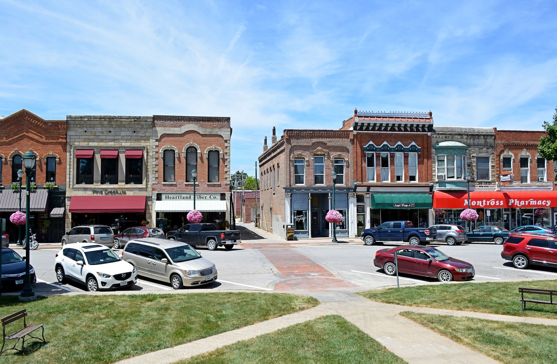 Downtown streetscape with brick buildings, shops, cars, and a green space on a sunny day.