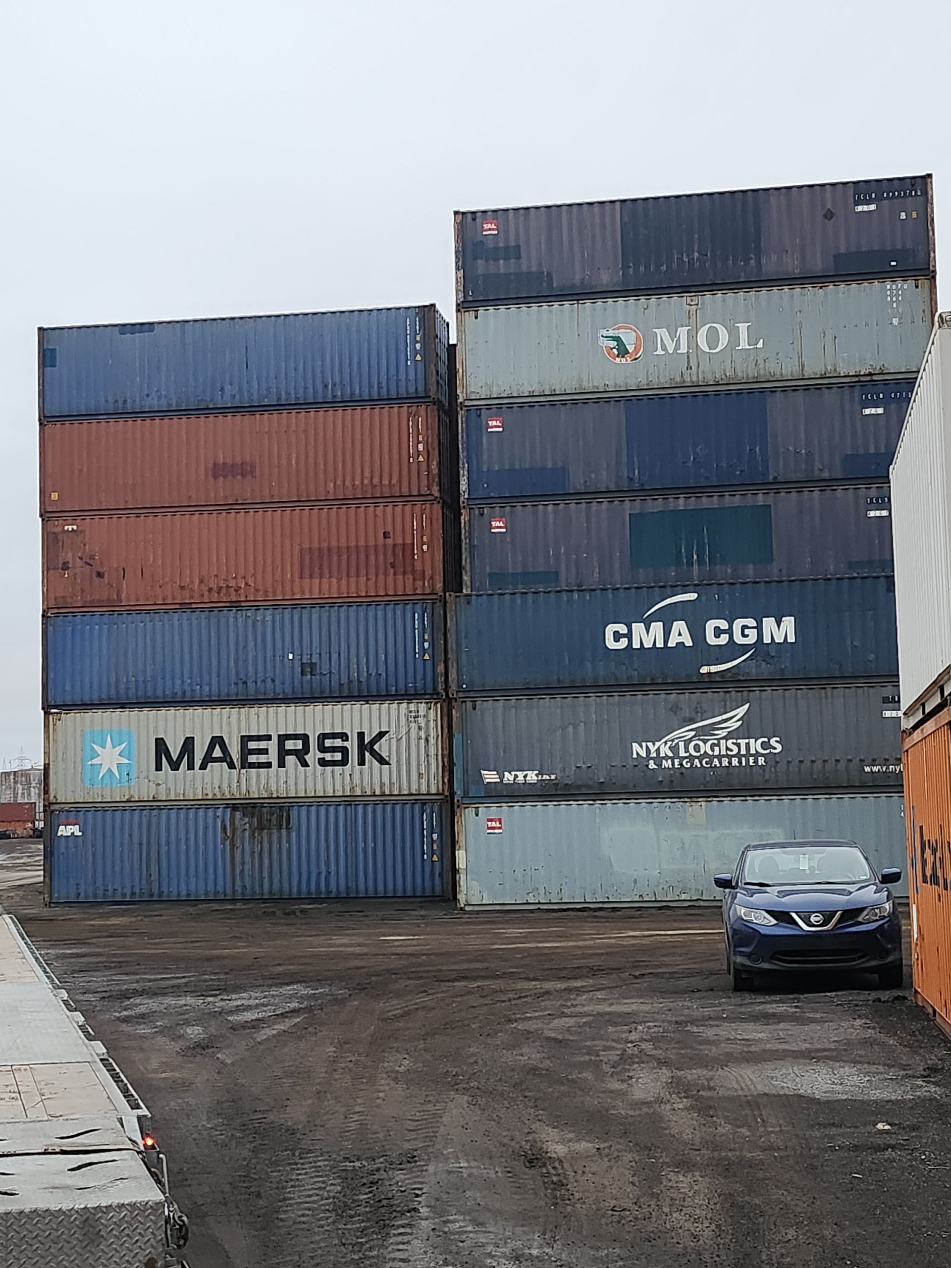 Une voiture est garée devant une pile de conteneurs maritimes.