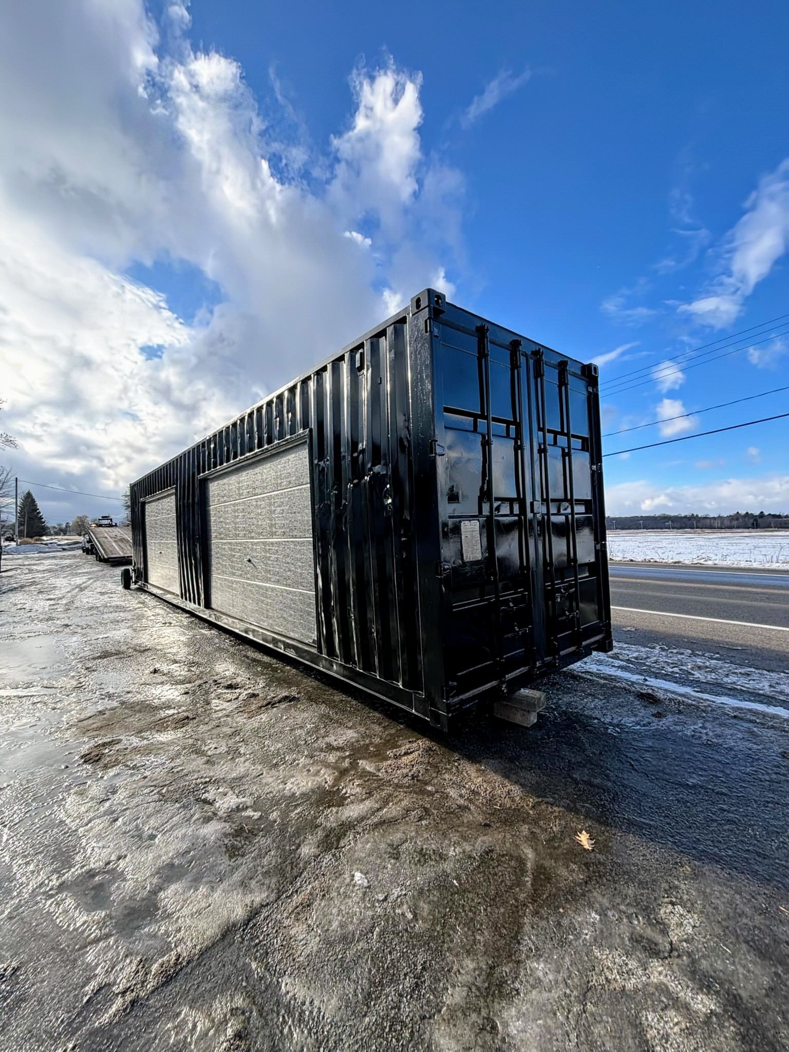 Un grand conteneur d'expédition noir est garé sur le bord de la route dans la neige.