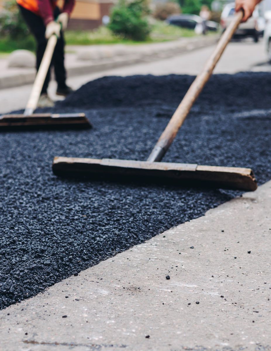 Workers smoothing fresh asphalt on a road with long-handled tools; close-up shows the dark black paving contrasted with a lighter-colored concrete edge.