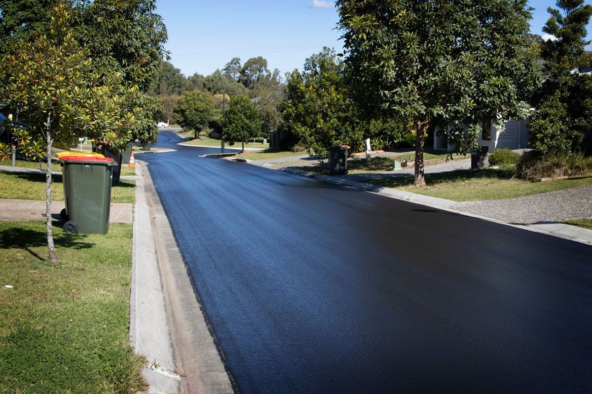 Newly paved residential street with trees, grass, and a green trash bin on a sunny day.