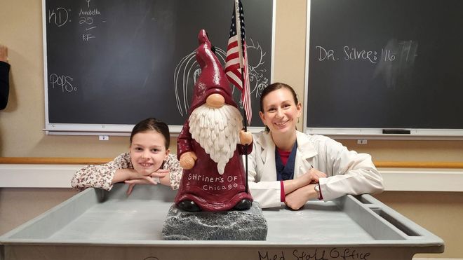 Two smiling females pose with a gnome statue in front of a chalkboard. The gnome holds an American flag.