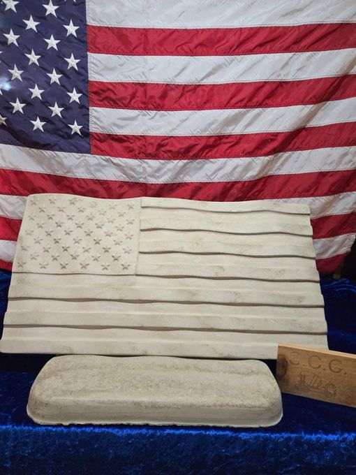American flag crafted from wood in front of a real American flag. A stone base and small brick are beneath the wooden flag.