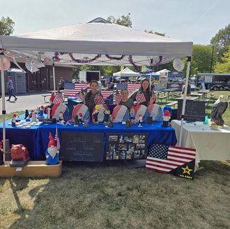 People at a craft fair booth decorated with American flags and patriotic merchandise. Three women hold flags; the table is blue.