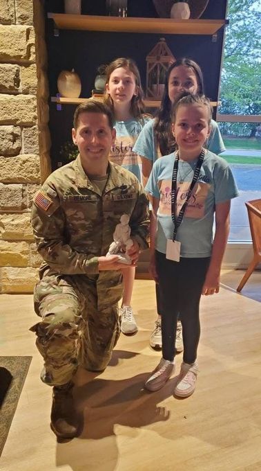 Soldier kneels, holding a statue, with three smiling girls in front of a shelf. Indoors with warm lighting and a window in the background.