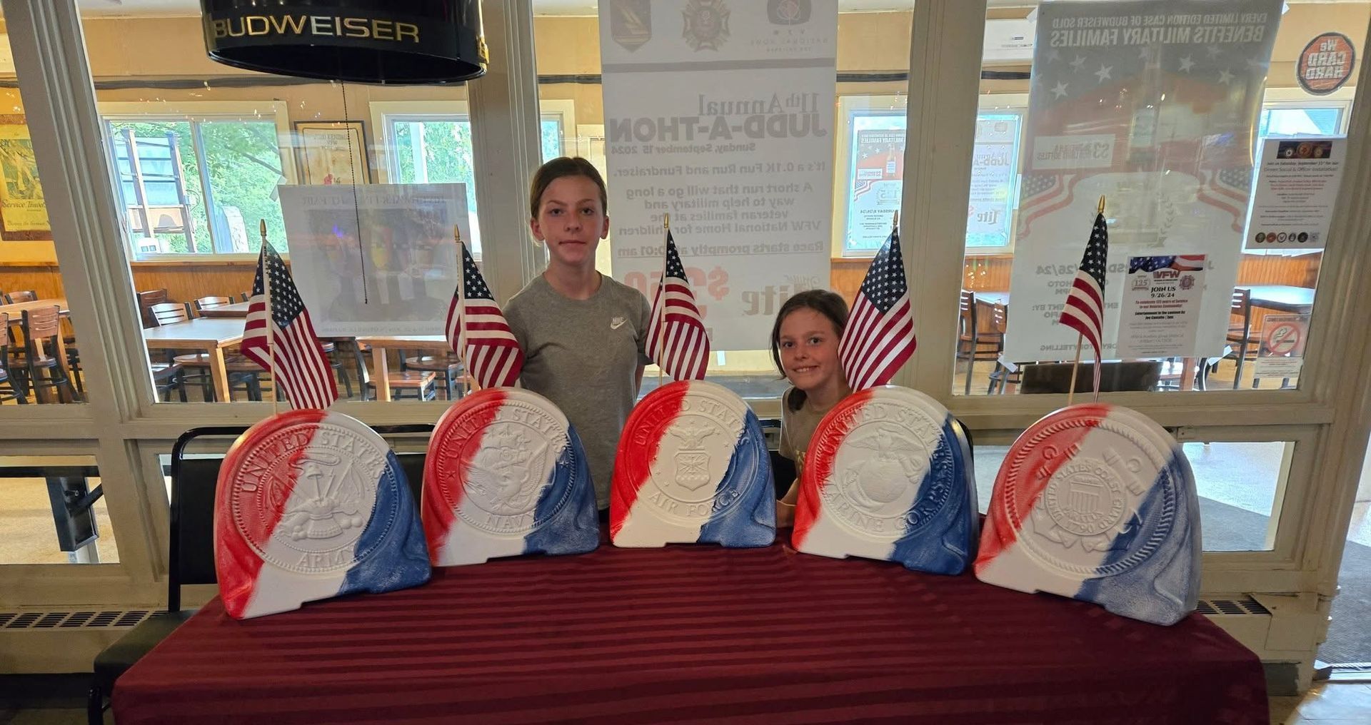 Two children pose behind patriotic art, red, white, and blue helmets, with American flags in a room with wooden accents.