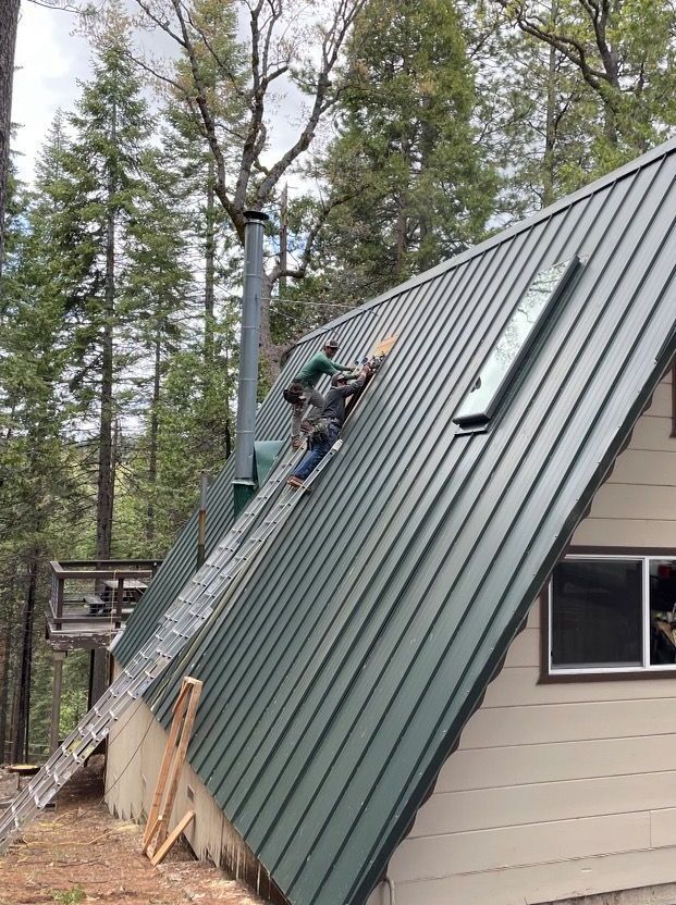 Person on a green metal roof installing wood trim. A-frame cabin with a chimney and skylight. Ladder.