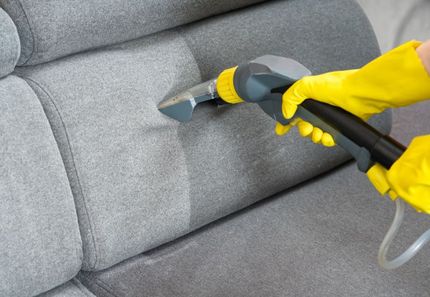 A person wearing yellow gloves is cleaning a couch with a vacuum cleaner.