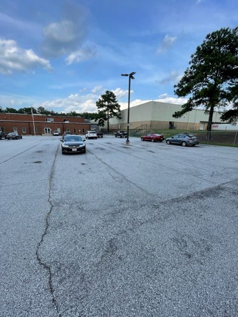 Cars parked in a cracked asphalt lot, under a blue sky. A building and trees are in the background.