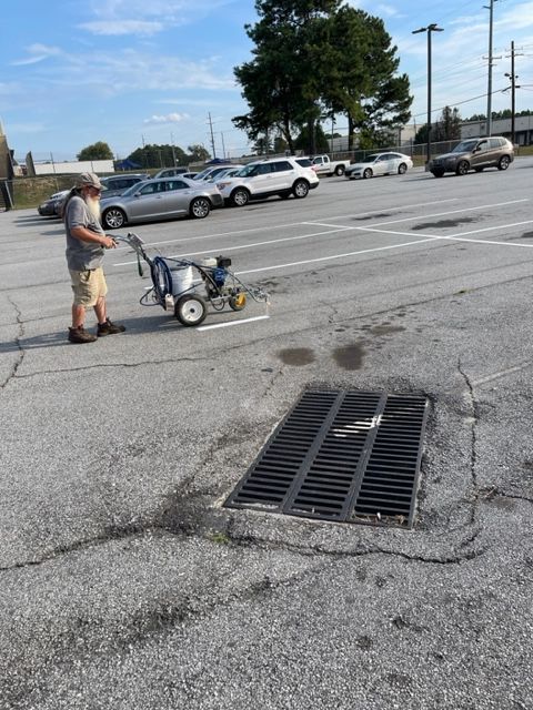 Man painting parking lines with a machine in a parking lot. Asphalt shows cracks near a storm drain, cars parked in background.