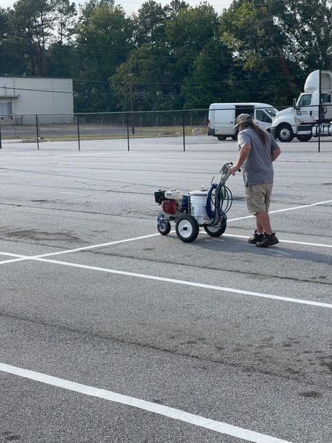 A person striping parking lines with a machine in a mostly empty parking lot. They are wearing shorts, a t-shirt and hat.