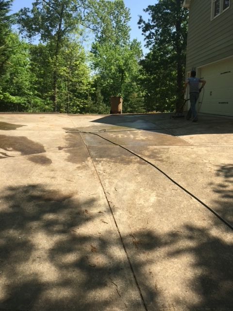 A person pressure washing a concrete driveway next to a house with a garage. Trees are in the background.