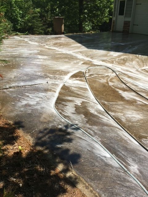 Driveway being cleaned with white foamy detergent, showing light and dark areas. A garden hose lies on the surface.