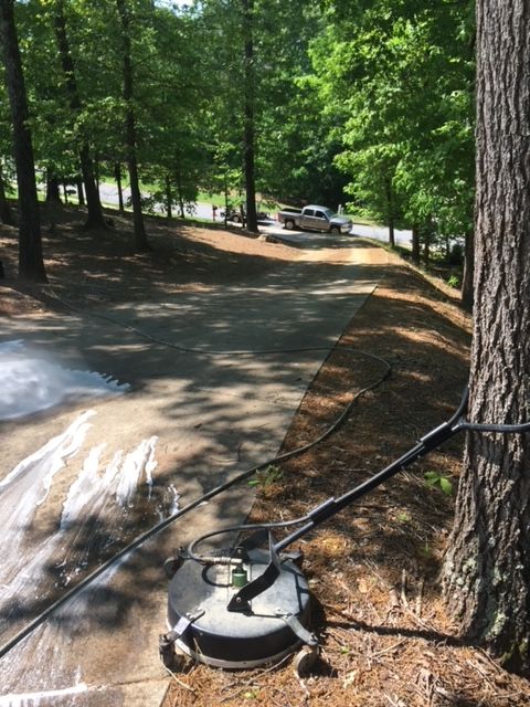 A person pressure washing a driveway, with a circular cleaning attachment. The driveway is surrounded by trees and grass on a sunny day.