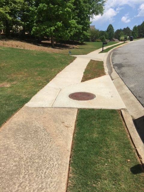 A sidewalk curves alongside a road, passing a manhole cover. Patches of grass border the concrete path under a partly cloudy sky.