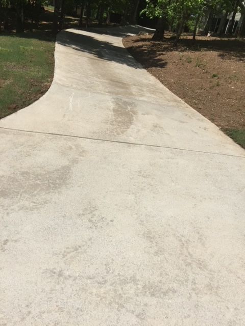 Concrete driveway curves towards trees, with patches of discoloration. Lush green grass and brown mulch border the sides.