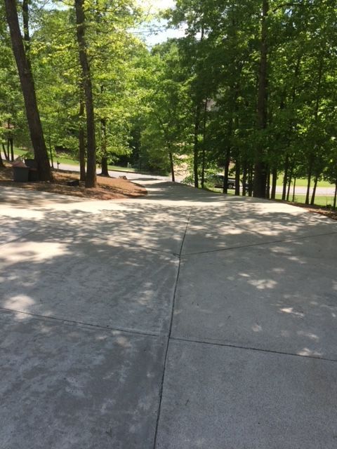 A paved driveway slopes downhill, surrounded by trees with green foliage in a sunny setting.