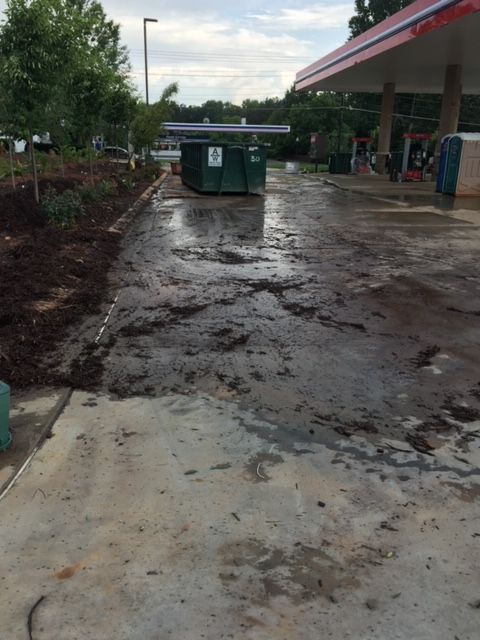 Muddy, wet concrete at a gas station. A green dumpster sits in the middle. Mulch borders a landscaped area.