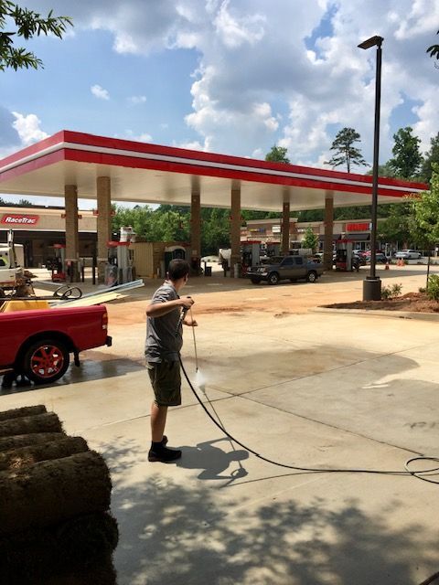 Man power-washing a gas station parking lot. Red and white canopy over gas pumps, red pickup truck, and a bright sunny day.