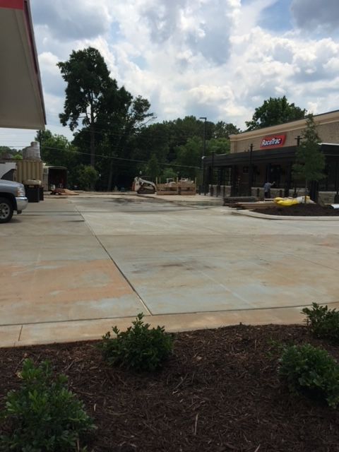 A wide shot of a mostly empty, light-colored concrete parking lot with a restaurant on the right and a gas station on the left under a cloudy sky.