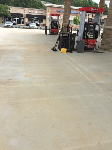 Gas station with pumps, a broom, and a trash can on a concrete surface. Retail stores are in the background.