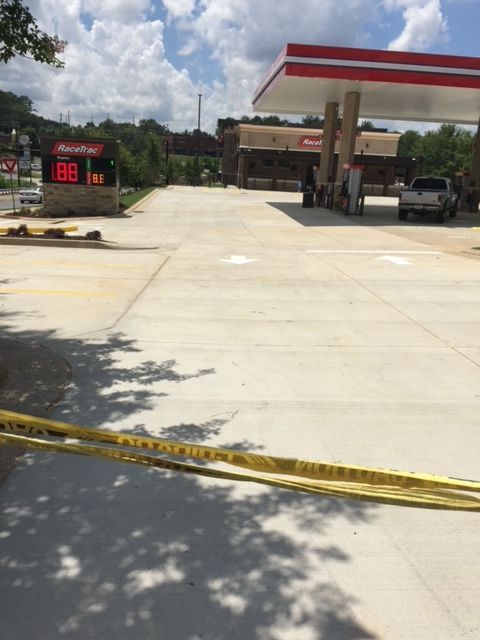 Gas station exterior with a canopy, pumps, and a price sign. Yellow caution tape blocks a concrete area under a bright, partly cloudy sky.