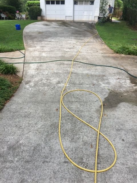Yellow hose coiled on a concrete driveway, leading towards a two-car garage. Green grass and bushes surround the driveway.