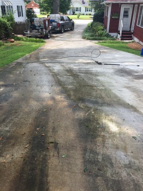 Driveway partially cleaned with a pressure washer, showing dark patches of dirt and lighter cleaned areas. A truck and surrounding houses are in the background.