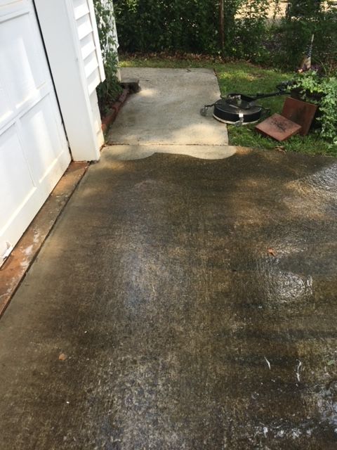 A person power washing a concrete walkway; showing before and after cleaning. The walkway is next to a white door, with surrounding green grass and foliage.