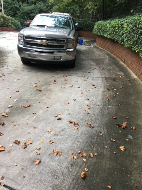 Silver pickup truck parked on a concrete driveway with fallen leaves. Green hedges and brick border the driveway.
