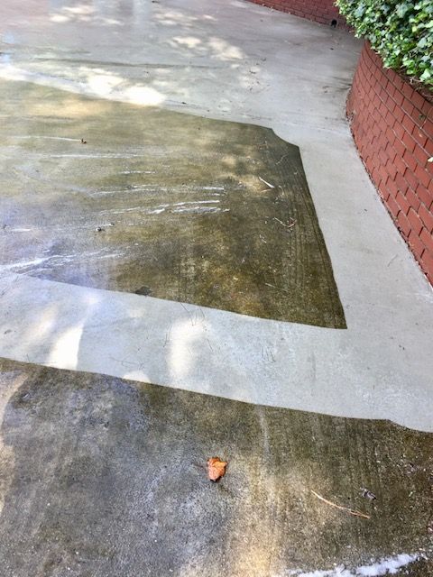 A concrete driveway, partially cleaned by a pressure washer, showing a distinct color difference. The edge of a red brick wall is in the right of the frame.