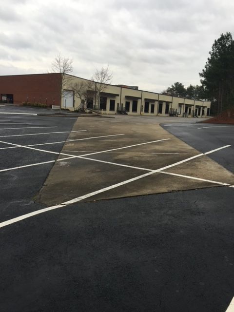 Empty parking lot in front of a one-story brick building with multiple storefronts. The pavement is dark gray with white parking lines.