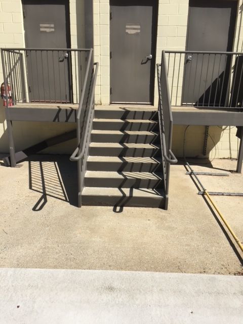 Exterior view of gray metal stairs leading up to three gray doors. Handrails and shadows are visible on a concrete platform.
