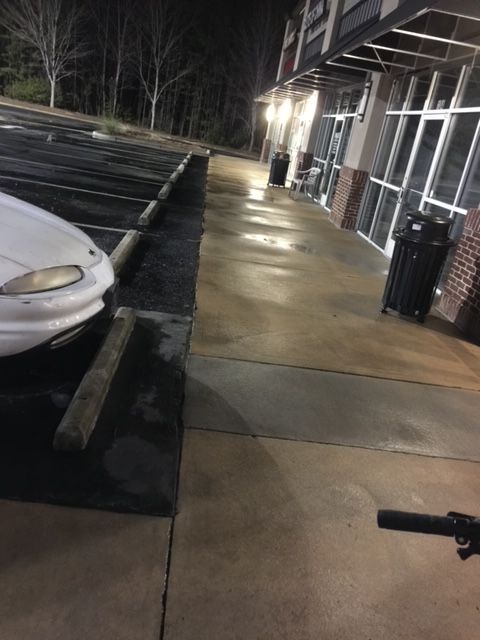 Nighttime view of a wet sidewalk next to a parking lot and a building with large windows. A white car is parked near the curb.