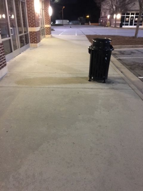 A black trash can on a concrete sidewalk outside a brick building, with an empty parking lot in the background at night.