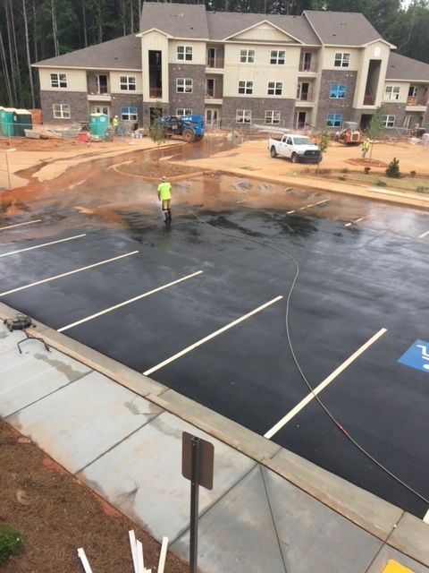 A person power washes a newly paved parking lot with marked spaces, in front of an apartment building under construction.
