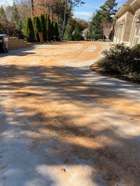 A concrete driveway covered in reddish-brown dirt, curving towards a brick house. Green trees and bushes line the edges.