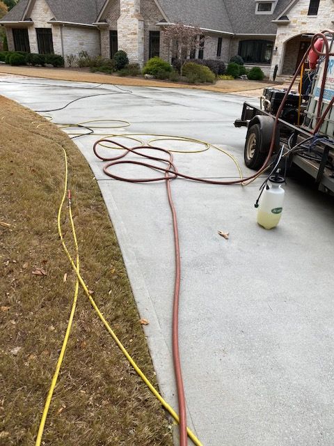 A concrete driveway being cleaned with a pressure washer. Hoses are laid out, and a trailer with equipment is parked on the side.