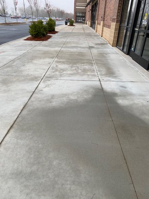 A concrete sidewalk in front of a building with windows and a door. There are small green bushes and a cloudy sky in the background.