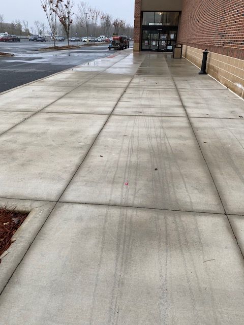 A concrete walkway leads to a store entrance. The sidewalk is wet, with a light gray color and a few leaves present.