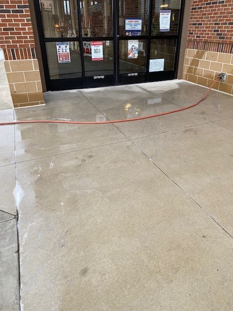 Wet concrete entryway with glass doors. A red hose lies on the wet surface. Brick and tan stone frame the doors.