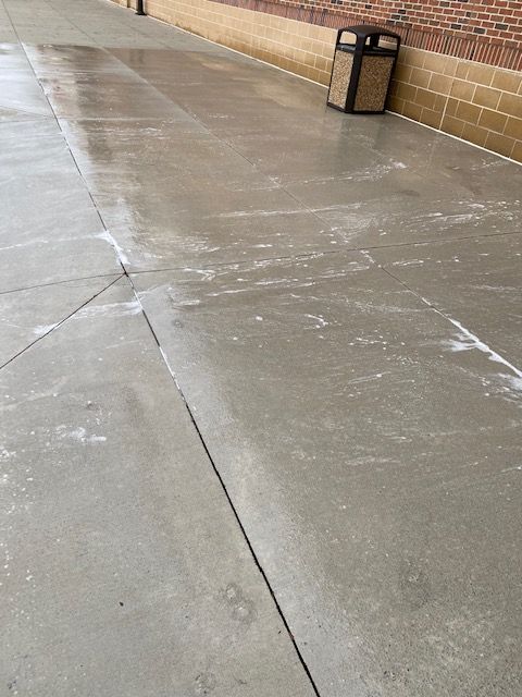 Wet concrete sidewalk with a trash can next to a brick building. Patches of snow or salt are visible on the surface.