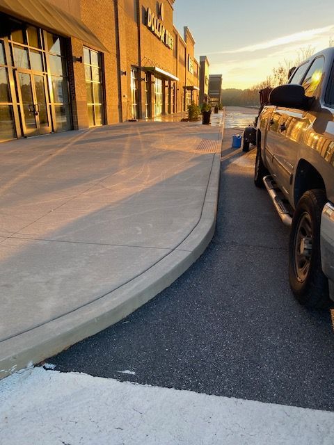 Sidewalk with curved curb next to an asphalt parking area, a truck is parked beside it. Golden sunlight bathes the scene near a brick building.
