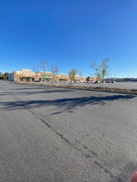 A large empty parking lot in front of a strip mall under a clear blue sky. The buildings are beige with sparse trees.
