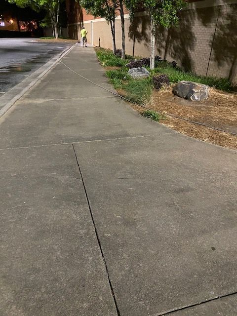 Sidewalk at night with a person walking in the distance. Trees, vegetation, and a brick wall line the right side.