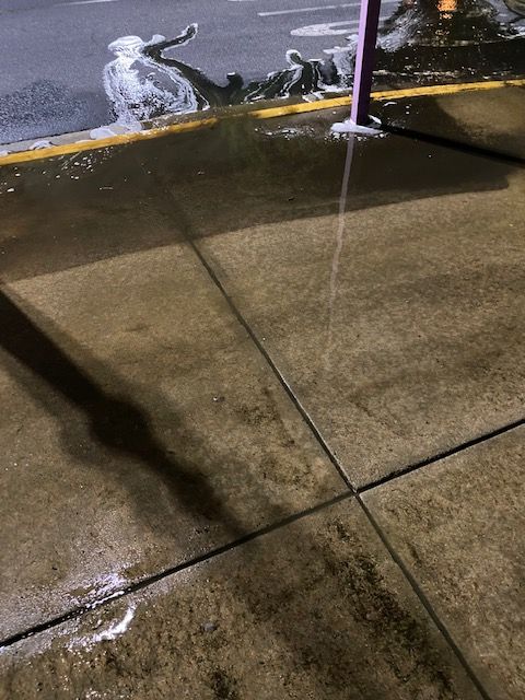 Wet concrete sidewalk with a long shadow cast across it; a purple post and some ice are visible in the background.