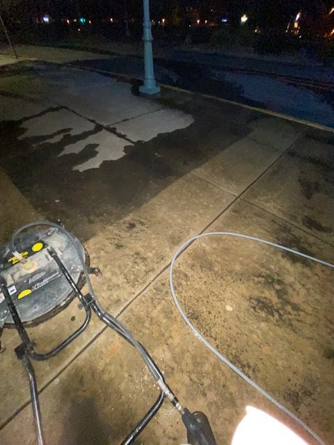 A pressure washer on a sidewalk at night, cleaning a dark, wet surface. A white cable and black hoses are visible.