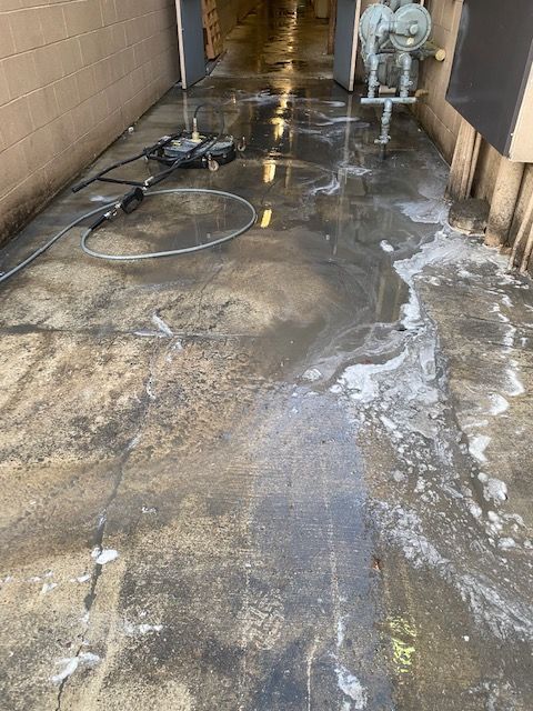 Concrete walkway being power washed, showing wet, dirty floor and equipment. Sunlight is entering from an open doorway.