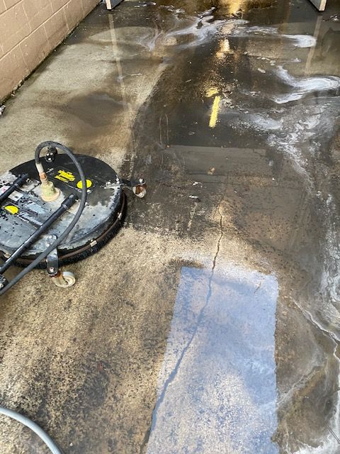 A pressure washer cleaning a concrete surface. Dark, wet area contrasts with dirtier, lighter areas.