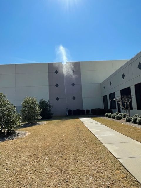 Water spraying from a warehouse wall on a sunny day. A long walkway extends toward the building.
